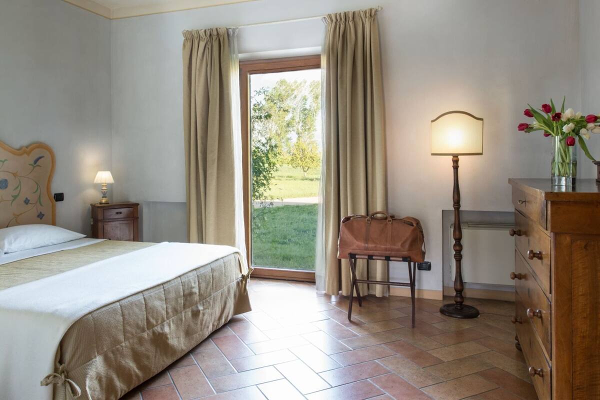 A hotel room with a bed, a nightstand, a lamp, a dresser, and a view of the outdoors.