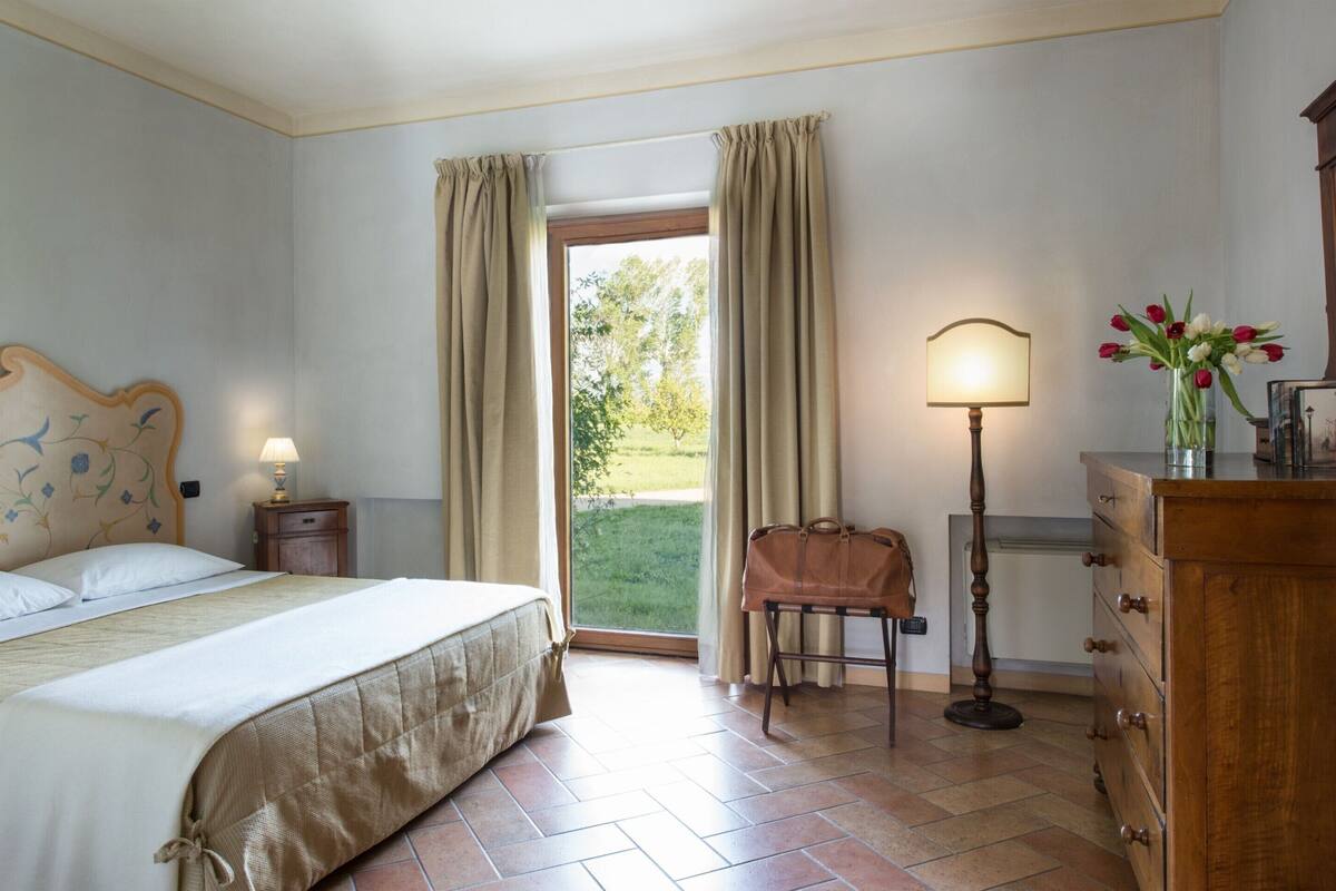 A hotel room with a bed, a nightstand, a lamp, a dresser, and a view of the outdoors.
