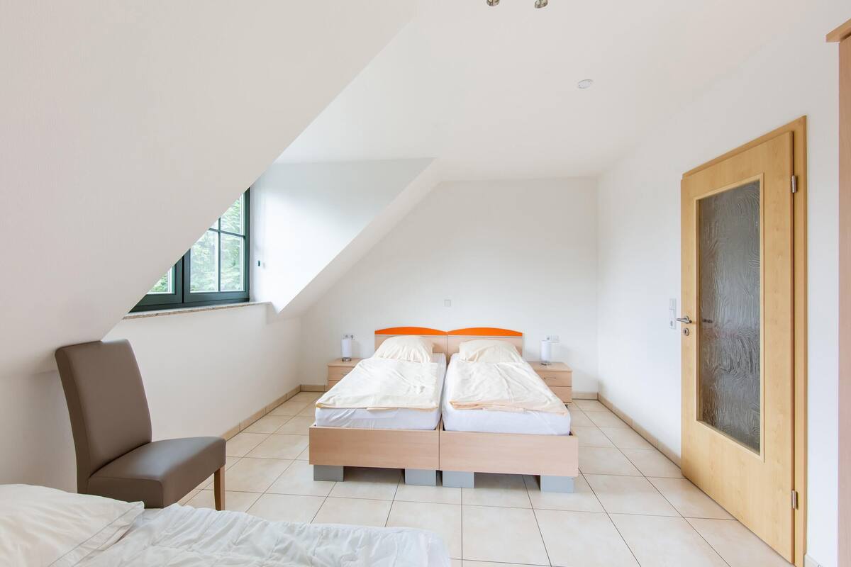 A bedroom with a sloped ceiling, two beds with orange headboards, a wooden chair, and a door with frosted glass.