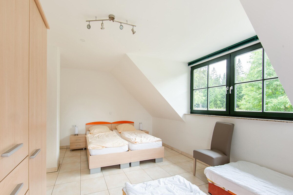 A bedroom with two beds, a chair, a wardrobe, and a window with a view of trees.