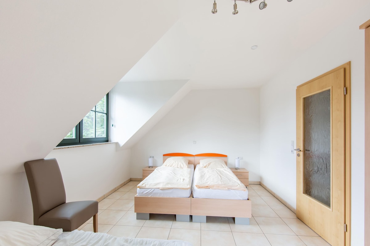 A bedroom with a sloped ceiling, two beds with orange headboards, a wooden chair, and a door with frosted glass.
