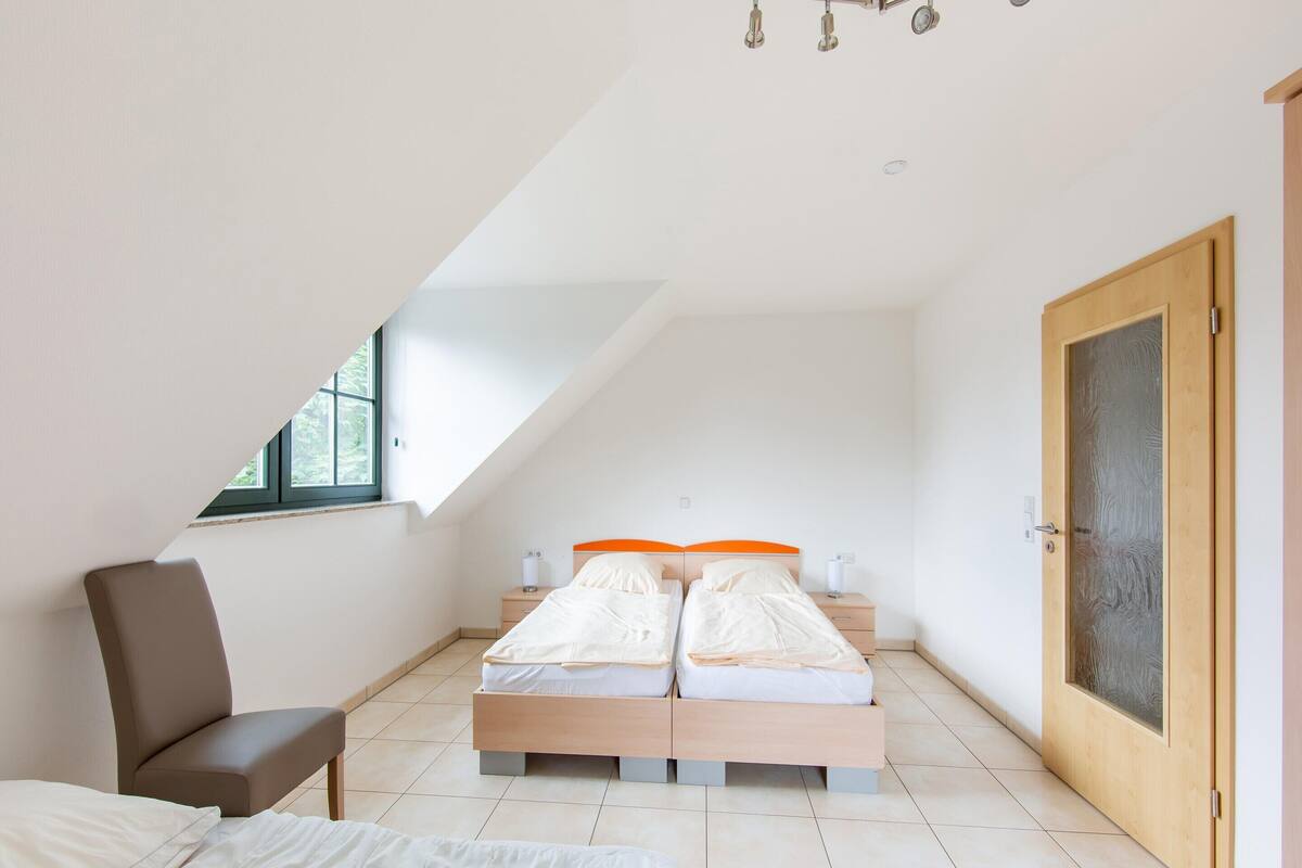 A bedroom with a sloped ceiling, a bed with orange headboards, a chair, and a wooden door.