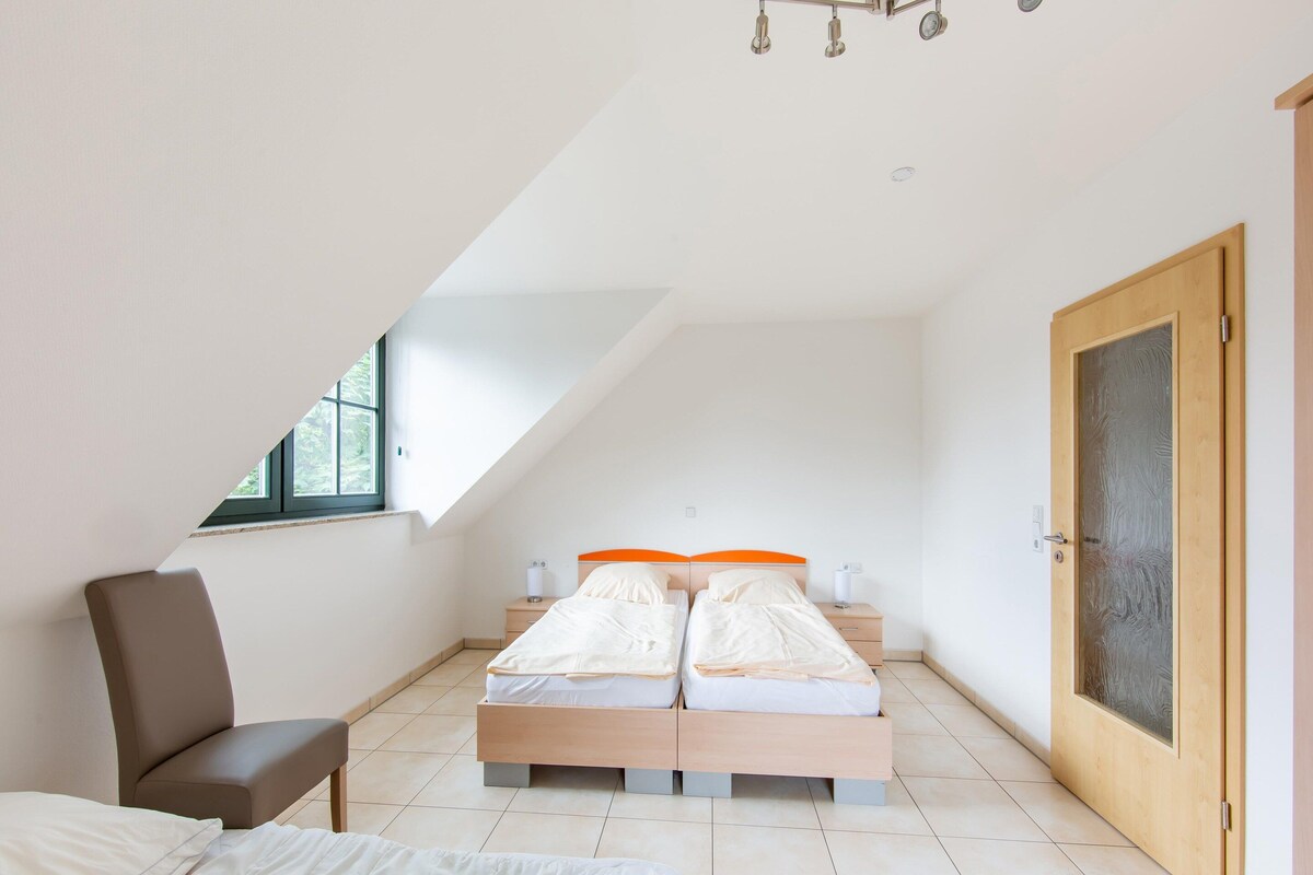 A bedroom with a sloped ceiling, a bed with orange headboards, a chair, and a wooden door.