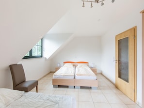 A bedroom with a sloped ceiling, two beds with orange headboards, a wooden chair, and a door with frosted glass. - Spacious Semi-detached House With Wood Stove Located Directly on the Rennsteig (Eisfeld)