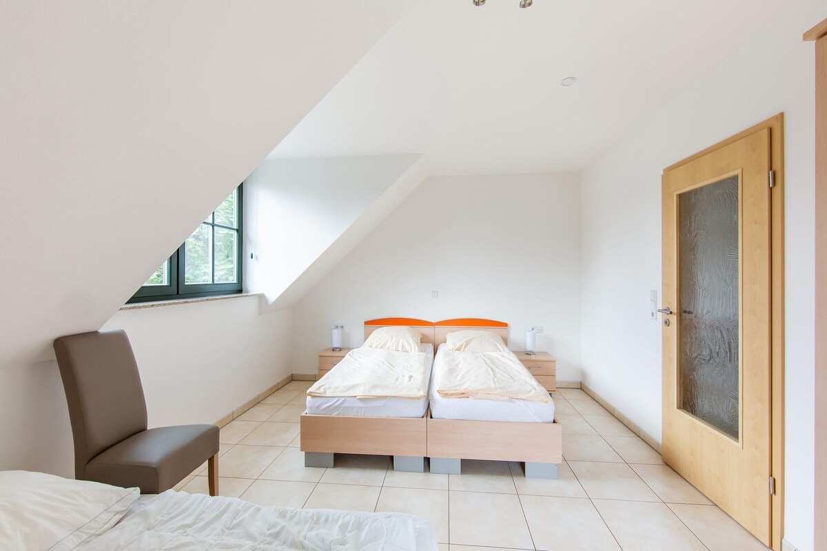 A bedroom with a sloped ceiling, two beds with orange headboards, a wooden chair, and a door with frosted glass.
