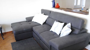 A grey sectional sofa with white and patterned cushions, a dark grey rug, and a tiled floor.