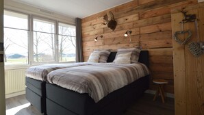 Un dormitorio con una cama grande, paneles de madera en la pared, un taburete pequeño y una ventana con vistas a árboles y edificios.