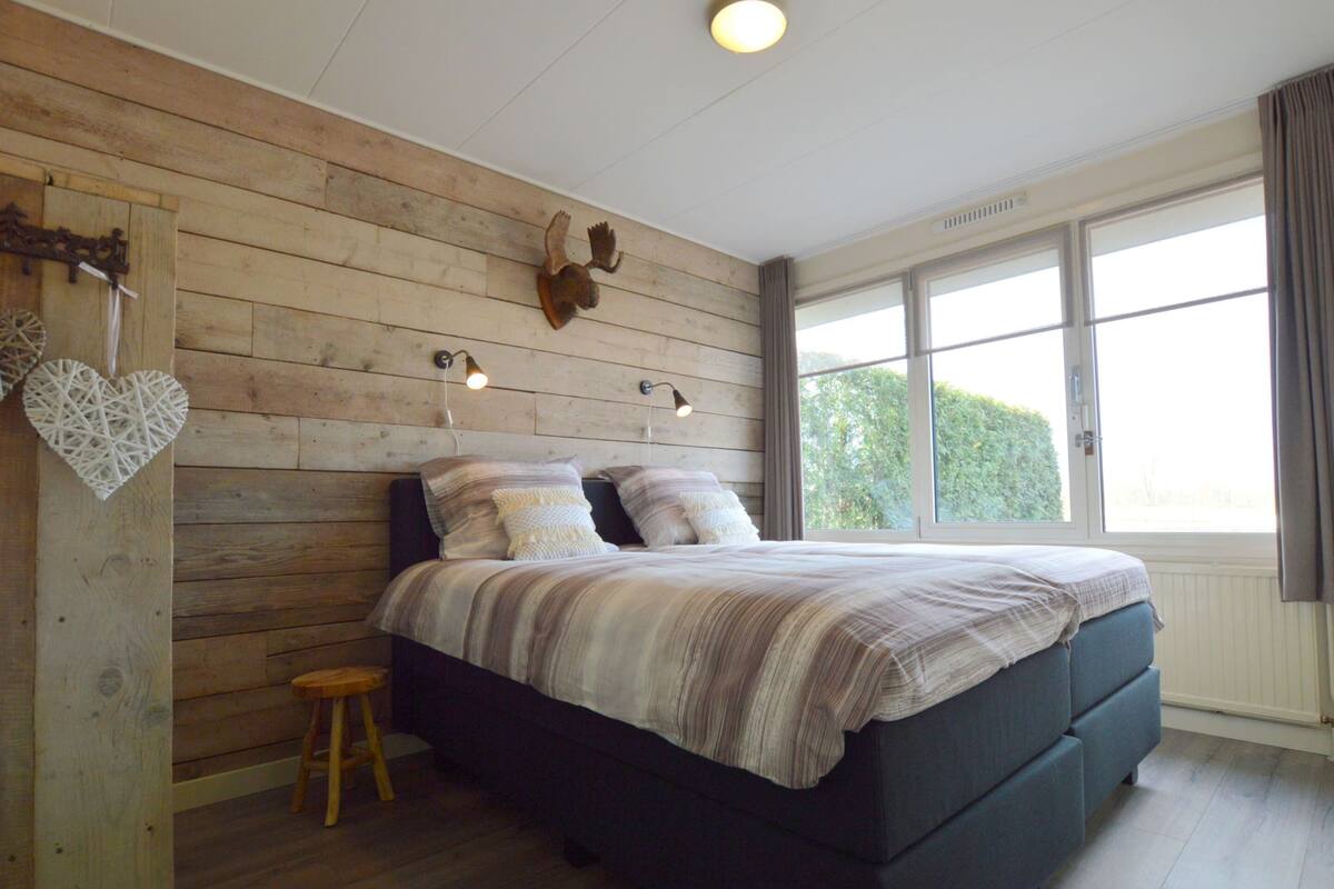 A bedroom with a wooden wall, a bed with a striped bedspread, a small stool, and a window with curtains.