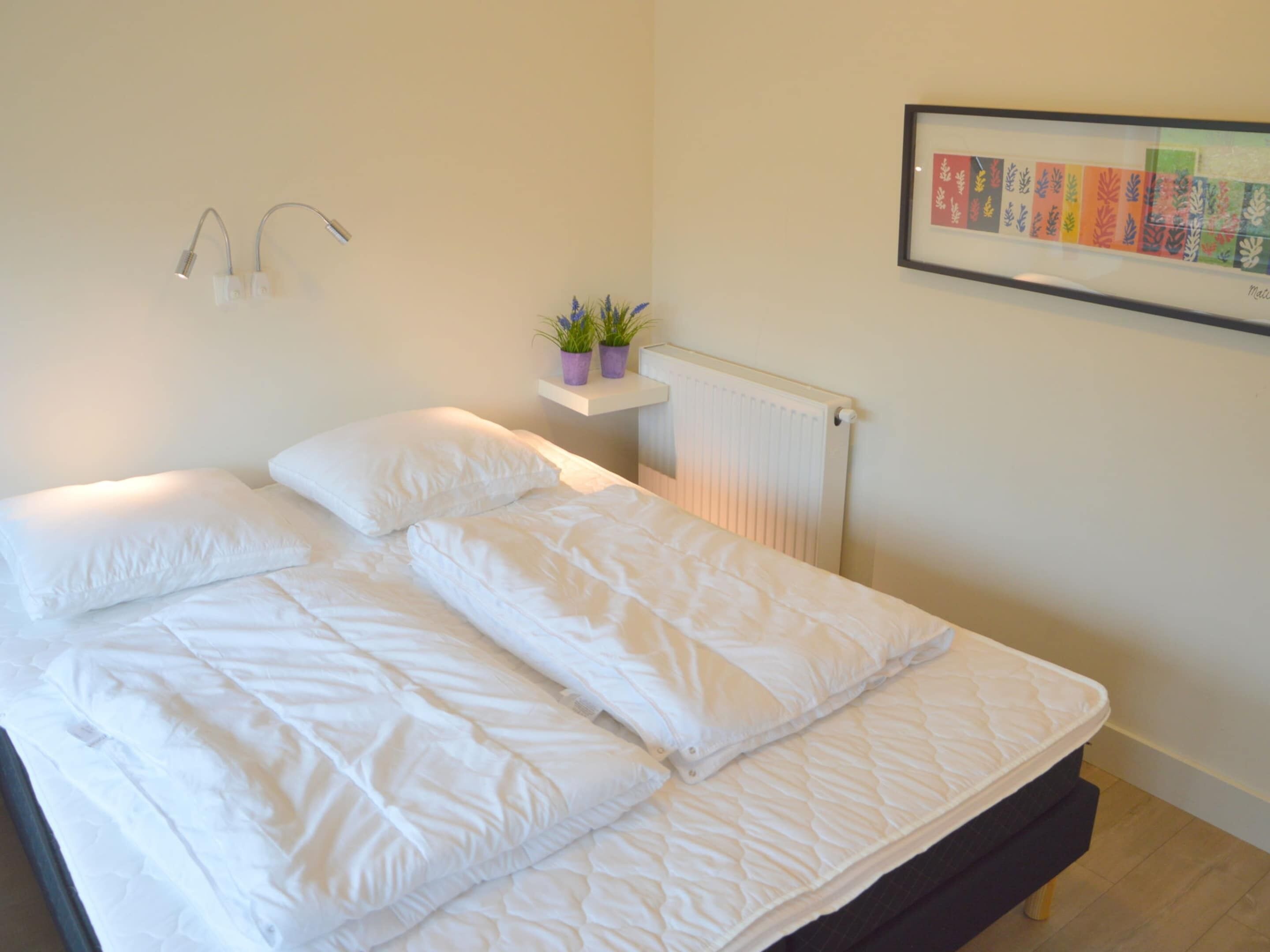 Una cama bien tendida con ropa de cama blanca, un radiador fijado a la pared y una estantería con plantas en macetas.