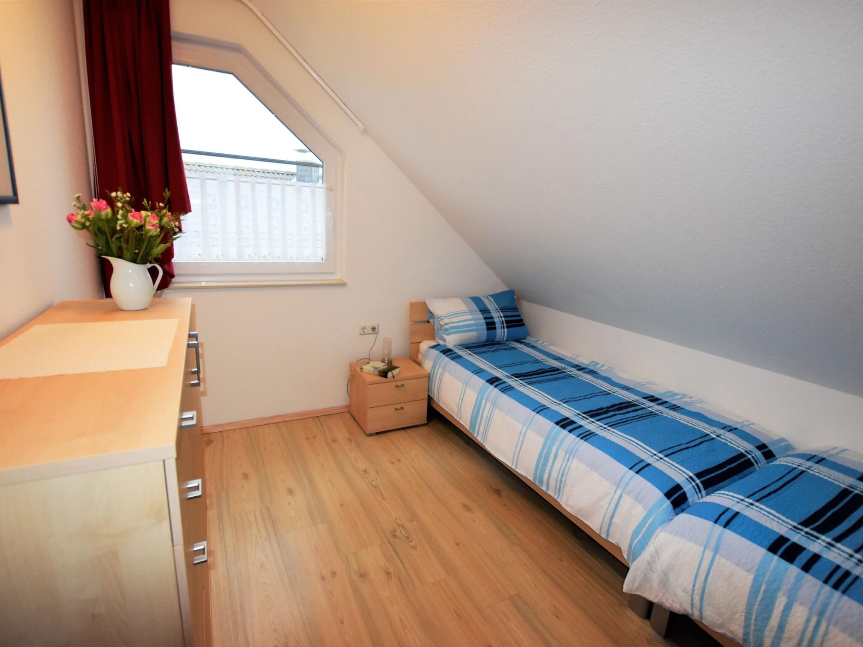 A small, tidy room with a bed, a nightstand, a wooden dresser, a window with curtains, and a vase of flowers.