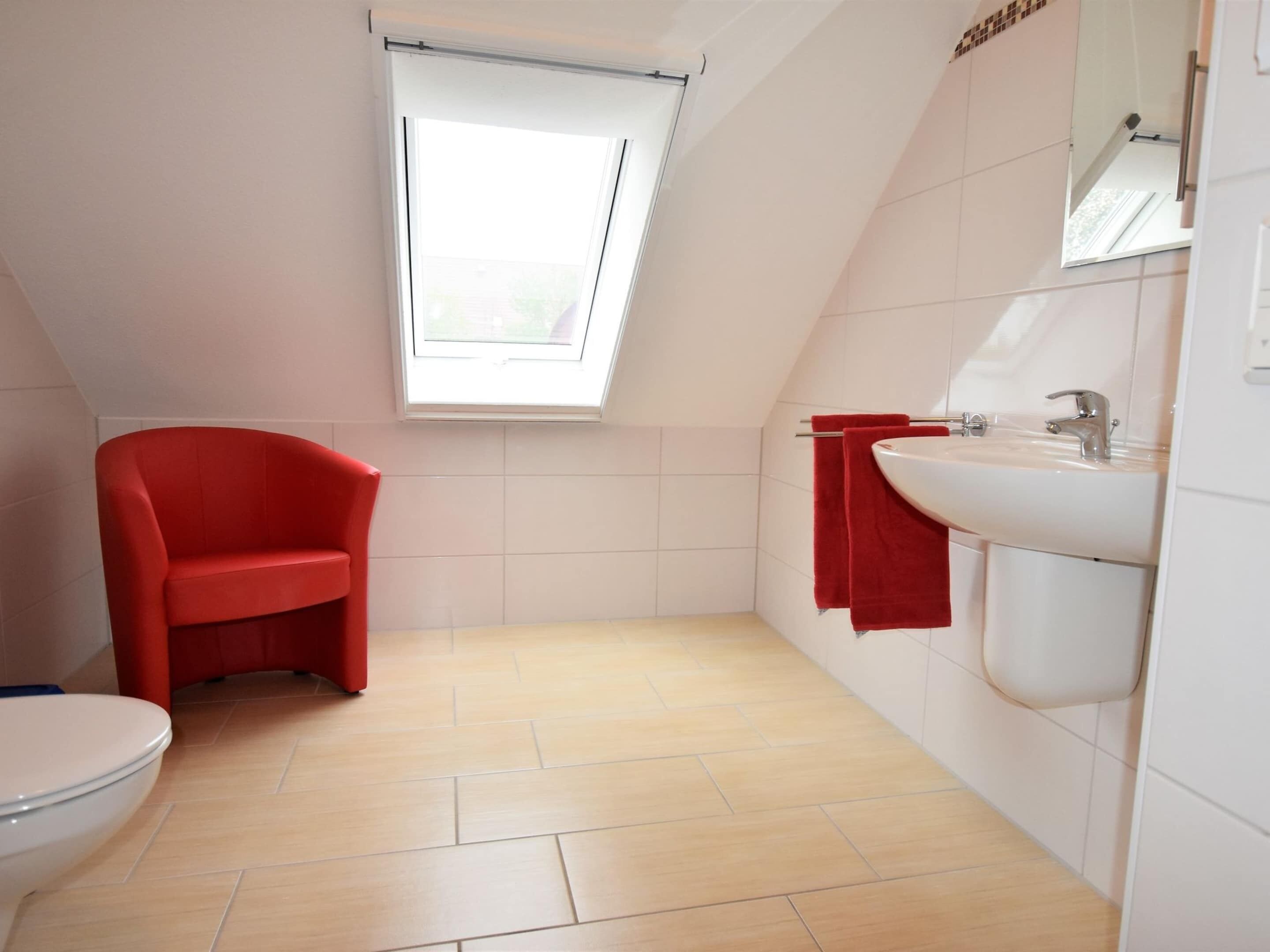A small, modern bathroom with a red chair, a toilet, a sink, and a window.