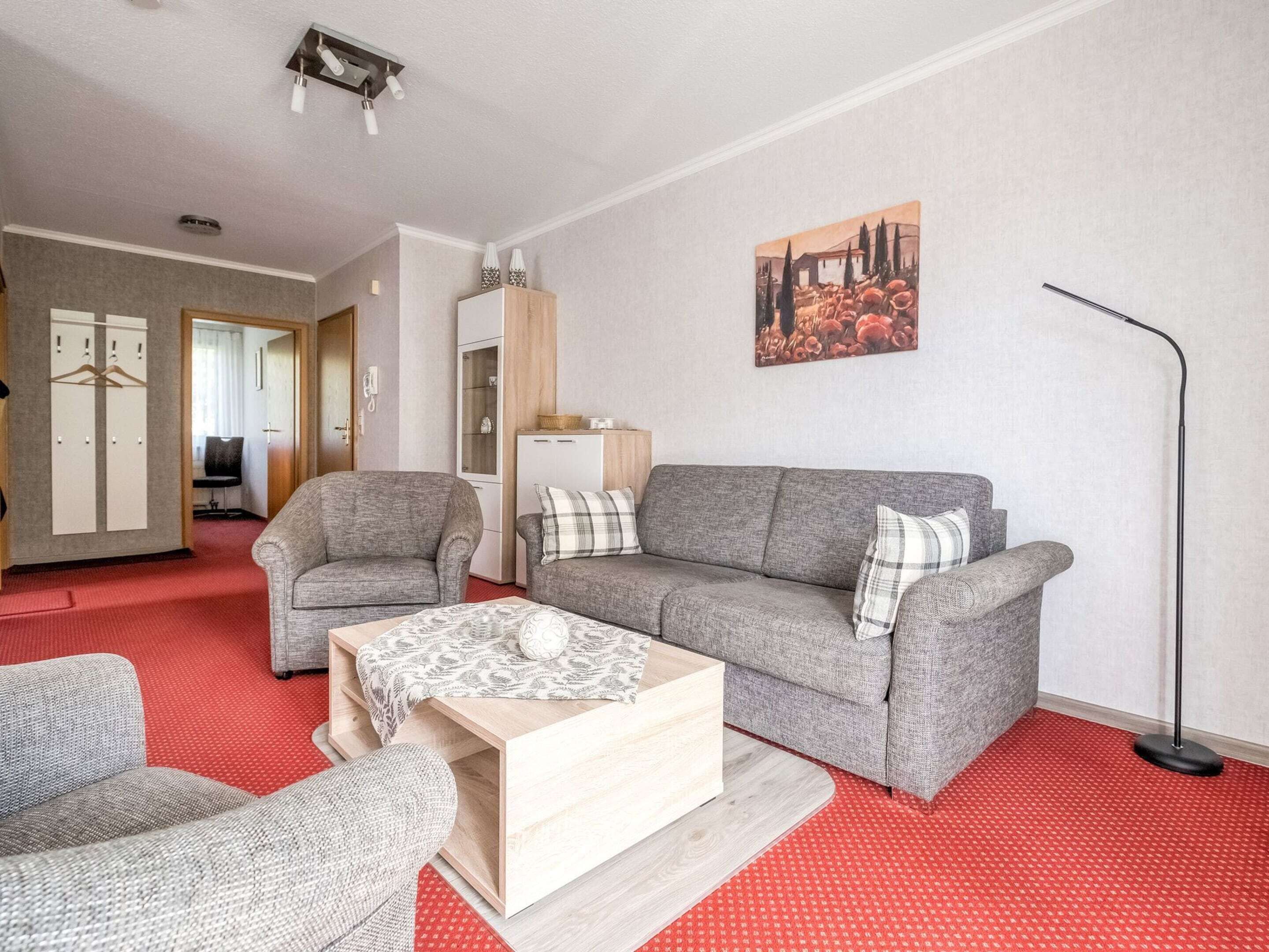 Un salon avec un canapé gris, deux fauteuils, une table basse et un tapis rouge. Il y a un tableau au mur et une lampe.