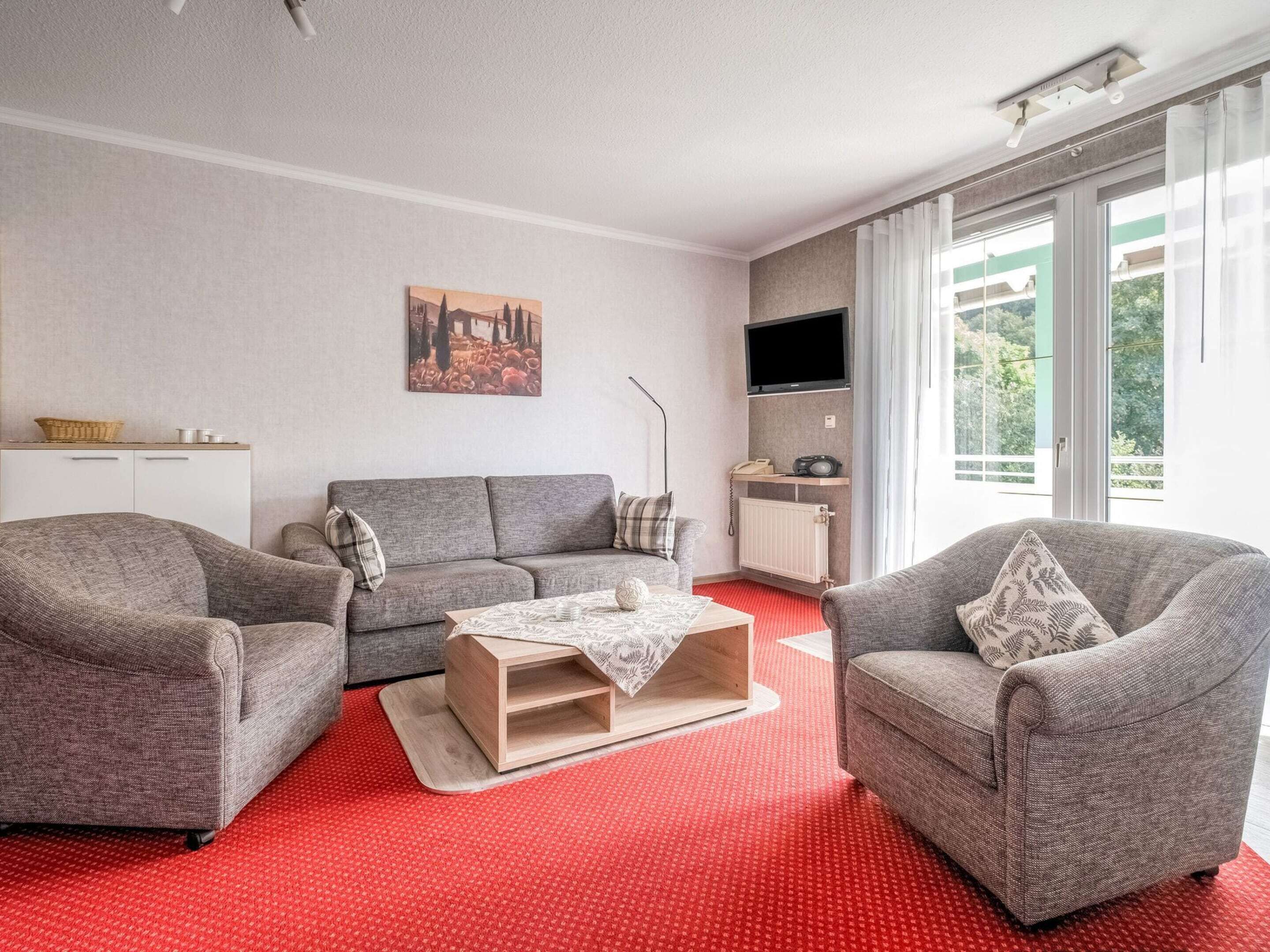 A living room with a grey sofa, two armchairs, a coffee table, and a red carpet. There is a painting on the wall, a TV, and a window with a view of greenery.