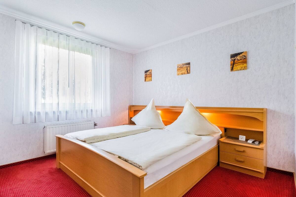 Une chambre d’hôtel avec un lit en bois, du linge de lit blanc, une moquette rouge, une table de chevet et deux tableaux accrochés au mur.