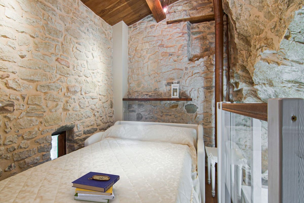 Quarto rústico com paredes de pedra, teto de madeira e cama com cobertura acolchoada. Há uma pequena estante com um candeeiro e uma pilha de livros na cama.