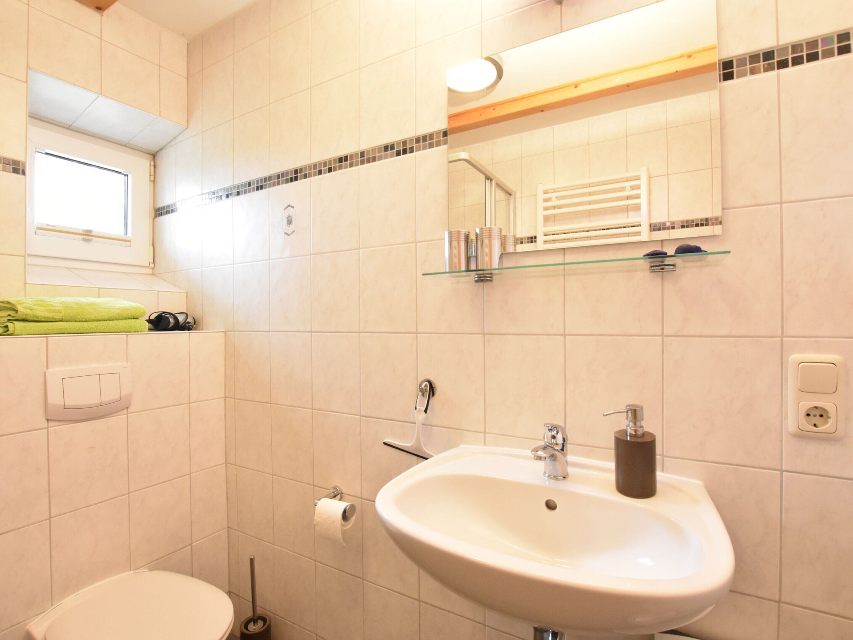 Egy fürdőszoba, amelyben fehér mosdókagyló, tükör, WC-papír-tartó és a mosdókagyló felett egy üvegpolc található.