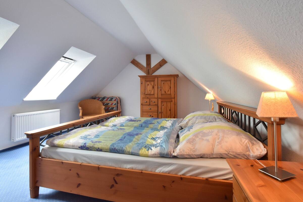 Ein Schlafzimmer mit schrägem Dach, einem hölzernen Bett, einem Sessel, einem Schrank und einer Lampe.