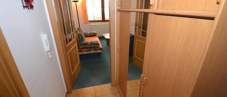 A narrow hallway with a wooden door, a small seating area with a colorful cushion, and a radiator in the background.