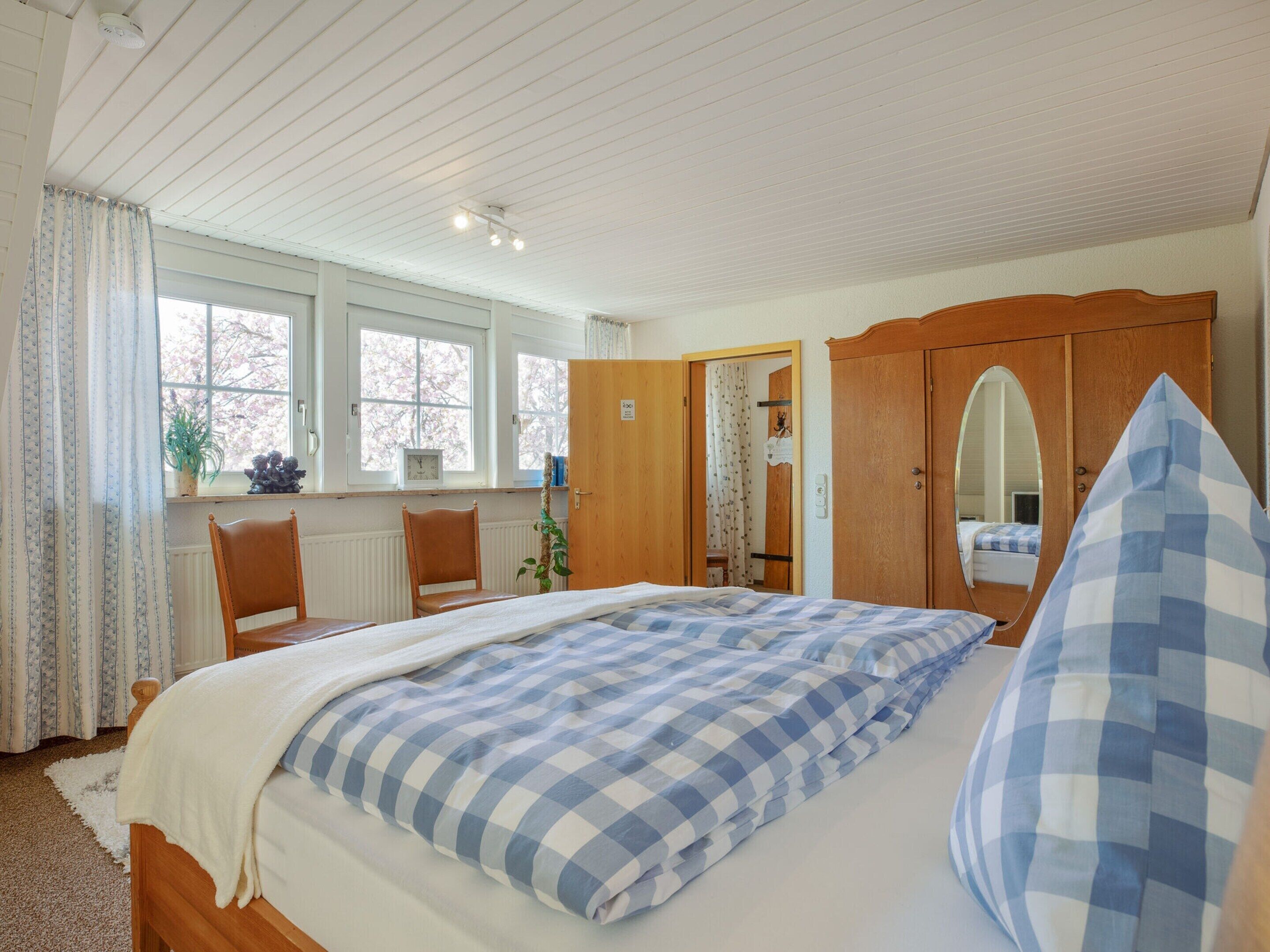 A bedroom with a bed, a wooden wardrobe, a chair, a window, and a mirror.