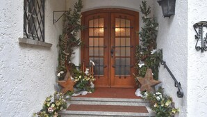 Une porte à deux battants en bois, ornée de panneaux de verre et flanquée de décorations en forme d’étoile et de verdure, qui mène à un escalier en pierre.