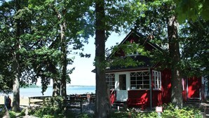 A red wooden building with a porch, surrounded by trees and a view of the water. - Lovely Holiday Home in Zierow With Terrace (Zierow)