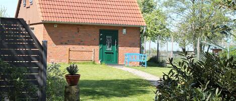 A brick building with a green door, a blue bench, and a garden with a tree stump and potted plant.