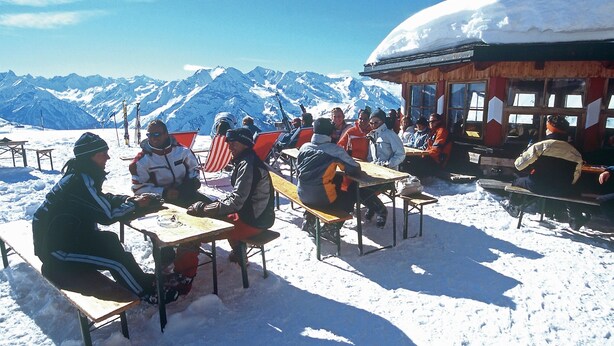 Un grupo de personas sentadas en mesas de madera al aire libre en un paisaje nevado, con montañas al fondo.