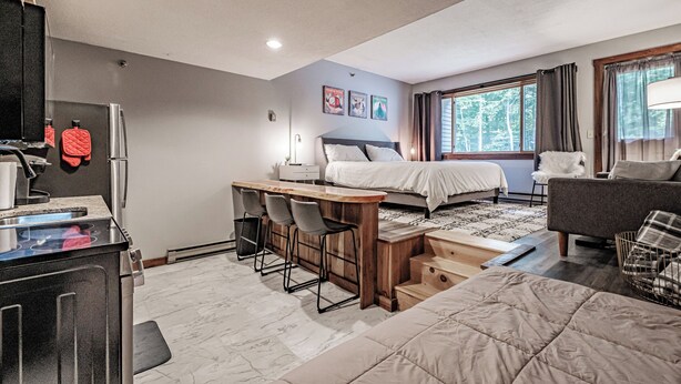 A modern hotel room with a bed, a desk with chairs, a sofa, and a window with curtains. - Cascades Studio at Sunday River (Newry)