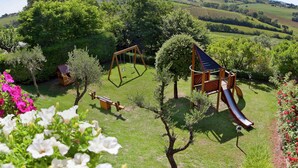 Um parquinho com balanços, escorrega e uma estrutura de escalada, cercado por árvores e flores.