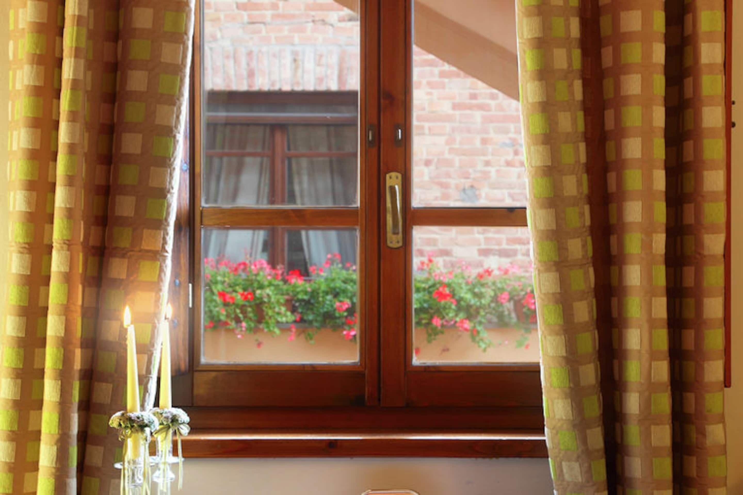 Een raam met houten kader en een messing handgreep, gordijnen met een geruit patroon, en een uitzicht op een bakstenen muur en rode bloemen buiten.