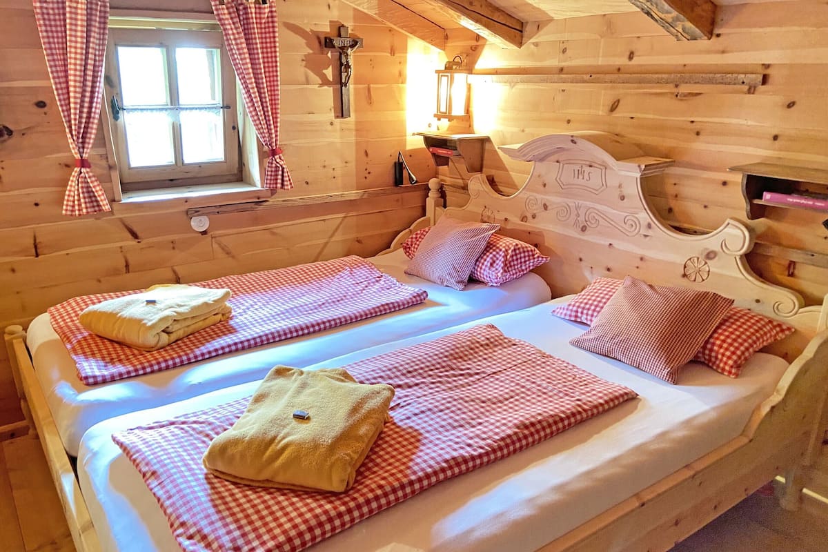 A wooden cabin room with two beds, checkered bedding, a window with curtains, and a cross on the wall.