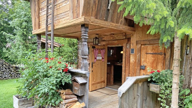Een rustiek houten gebouw met een houten trap die naar een veranda leidt, omringd door groen en een stapel hout voor de kachel.