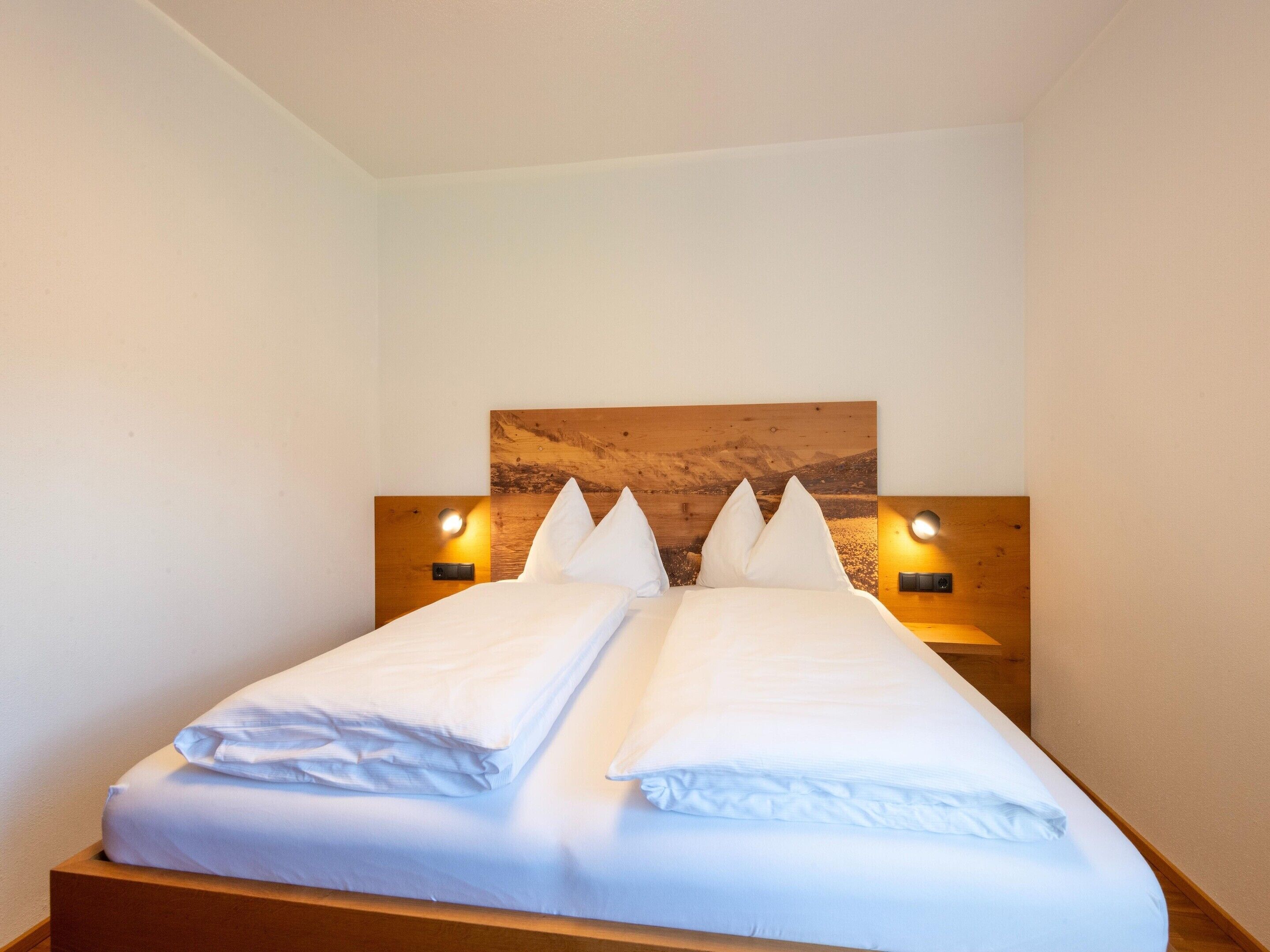 Un lit double avec du linge de lit blanc, une tête de lit en bois et deux lampes de lecture fixées au mur.
