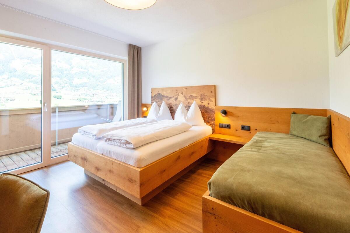 Ein Hotelzimmer mit einem großen Bett, einem Einzelbett, einem hölzernen Kopfteil, einem Nachttisch und Blick ins Freie durch eine Schiebetür.