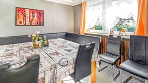 A dining area with a table set for four, a vase with flowers, and a bottle of champagne. There is a window with curtains, a painting on the wall, and a view of the outside. - Ferienhaus in Medebach Nahe Skigebiet (Medebach)