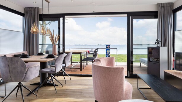 A modern dining area with a view of the sea, featuring a wooden table, grey and pink chairs, and a television on a cabinet. - Unique Houseboat on and Around the Sneekermeer (Offingawier)