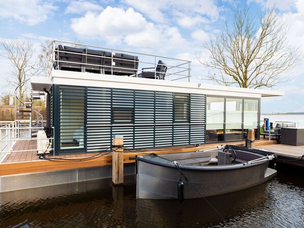 Une maison flottante moderne dotée d’une terrasse, avec un petit bateau amarré devant, et offrant une vue sur l’eau.