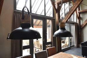 Un comedor con una mesa y sillas de madera, ventanas grandes con cortinas y lámparas colgantes negras.