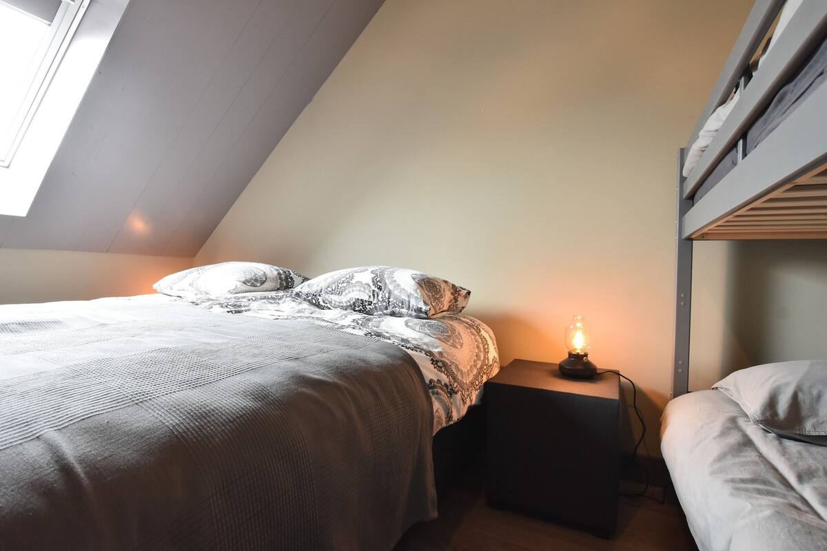 A bedroom with a bunk bed, a bedside table with a lamp, and a window providing natural light.