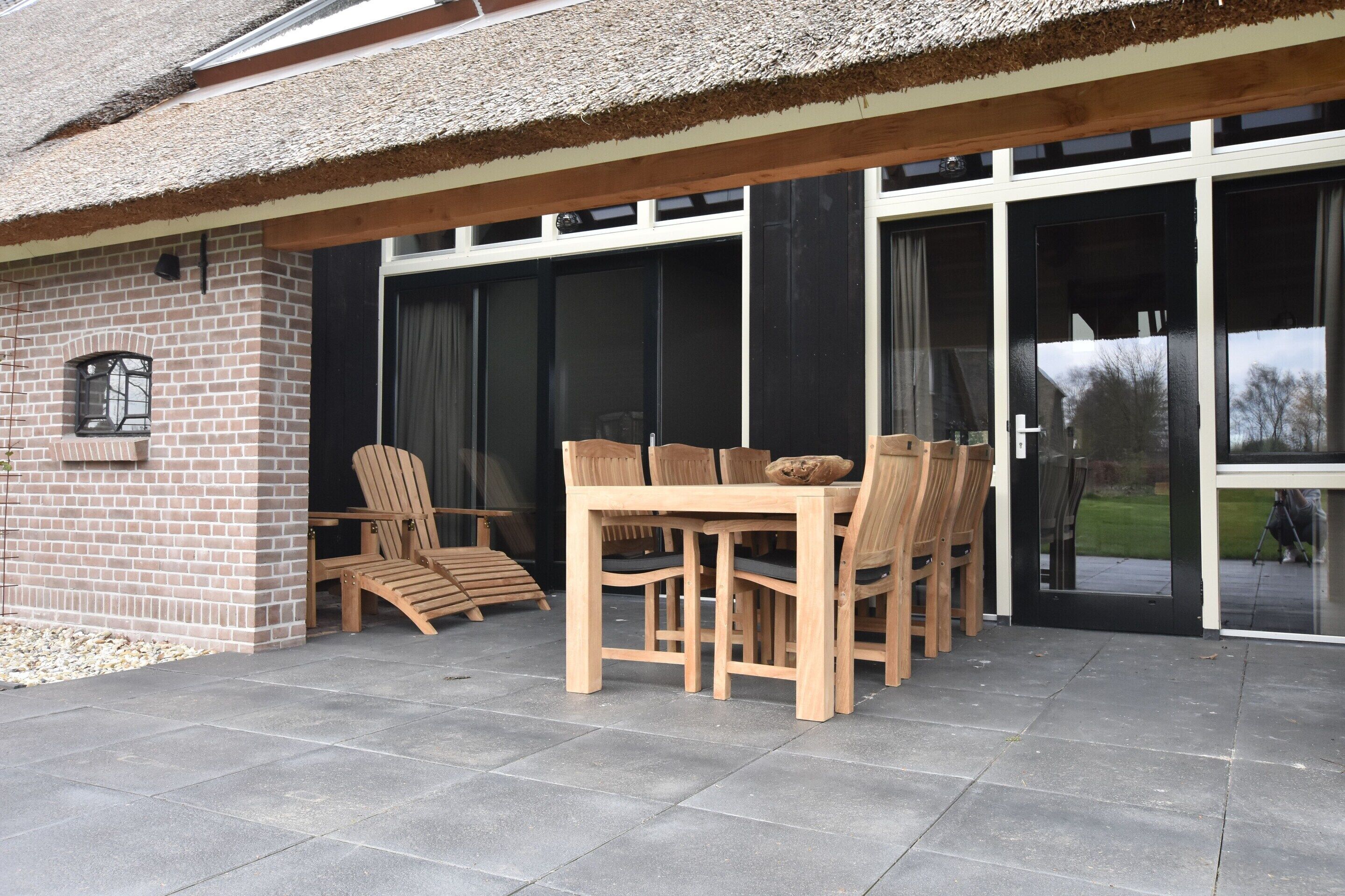 Eine Terrasse mit Backsteinmauer, Holztisch und -stühlen sowie einer Glastür, die in einen Garten führt.