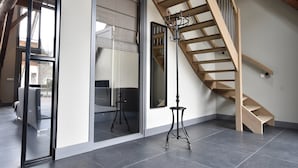 Un interior moderno con una escalera de madera, un vidrio que da a un balcón y una pequeña mesa con una lámpara.