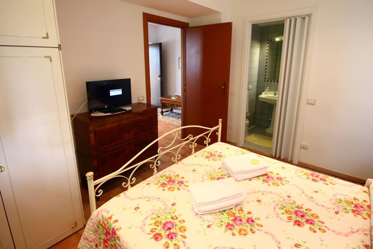 Une chambre à coucher avec un lit, une commode, un téléviseur et une salle de bain visible à travers une porte ouverte.