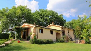 Una casa a due piani con tetto in tegole, circondata da vegetazione lussureggiante e alberi.