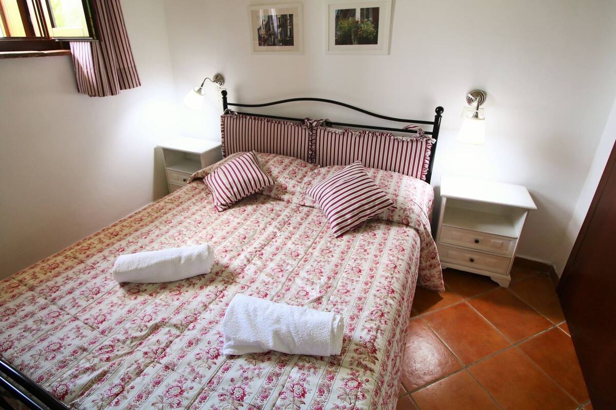 Un lit bien fait, avec une literie à motifs floraux, deux oreillers et deux serviettes. On y trouve deux tables de chevet avec des lampes, une petite table d’appoint et un tableau accroché au mur.