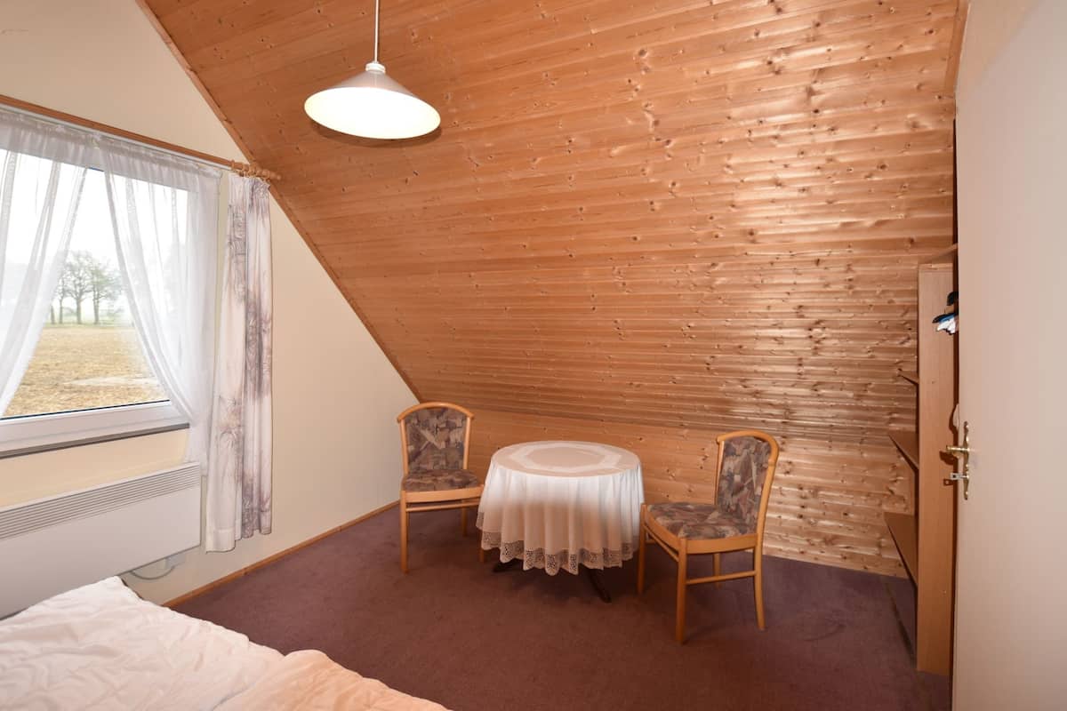 A room with a sloped ceiling, a bed, a table with chairs, a window with curtains, and a door.