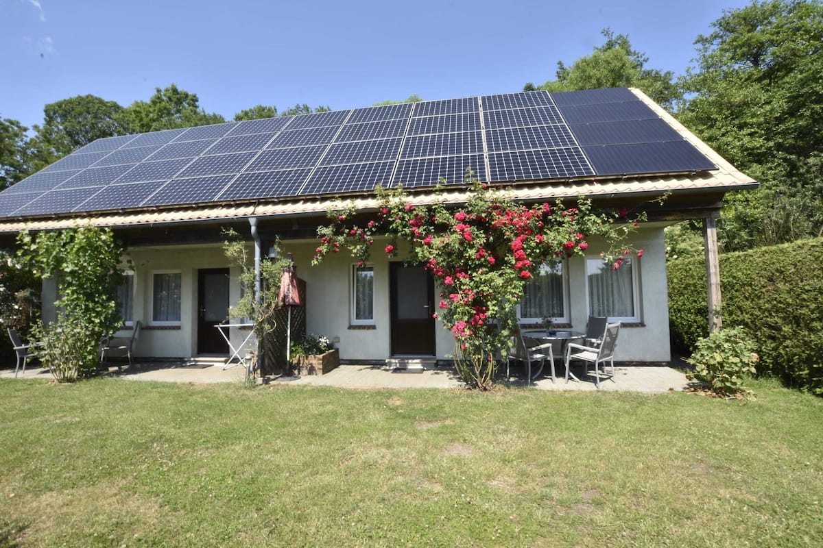 A house with a solar panel roof, a patio with chairs, and a garden with pink flowers.