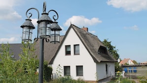 Une maison blanche avec un toit de chaume, un lampadaire en métal noir et un petit jardin avec des plantes et une chaise.