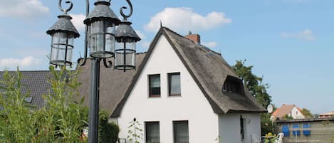 A white house with a thatched roof, a black metal lamp post, and a small garden with plants and a chair.