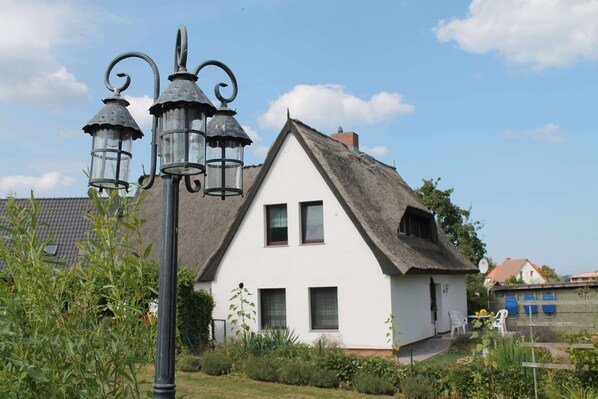A white house with a thatched roof, a black metal lamp post, and a small garden with plants and a chair. - Cosy Apartment in Pepelow Near Baltic Sea (Am Salzhaff)