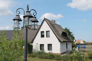 Uma casa branca com telhado de palha, um poste de luz de metal preto e um pequeno jardim com plantas e uma cadeira.